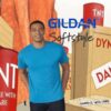 64000 Male model wearing a bright blue Gildan Softstyle fitted t-shirt against a cartoon-style background with dynamite crates and TNT barrels