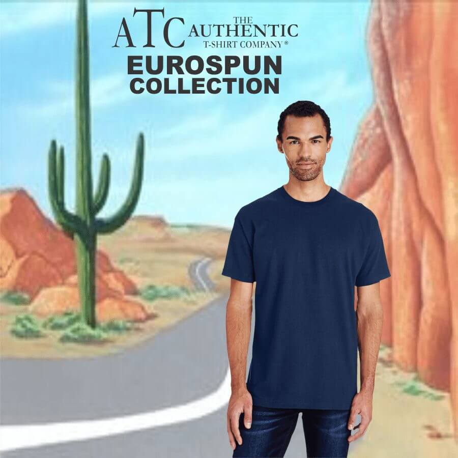 Male model wearing a navy blue short-sleeve crew neck t-shirt from ATC Authentic T-Shirt Company's Eurospun Collection, standing in front of a desert road backdrop with cactus and red rock formations