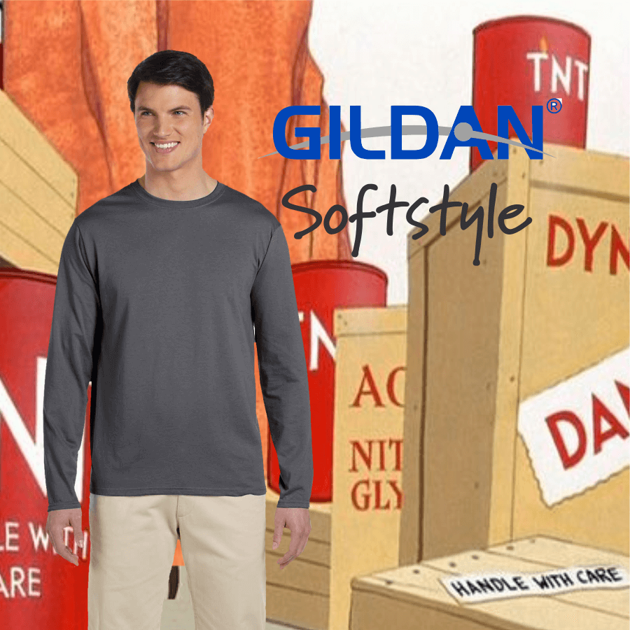 Male model wearing a dark gray long-sleeve Gildan Softstyle T-shirt paired with beige pants, standing against a cartoon background featuring wooden crates and red TNT barrels.