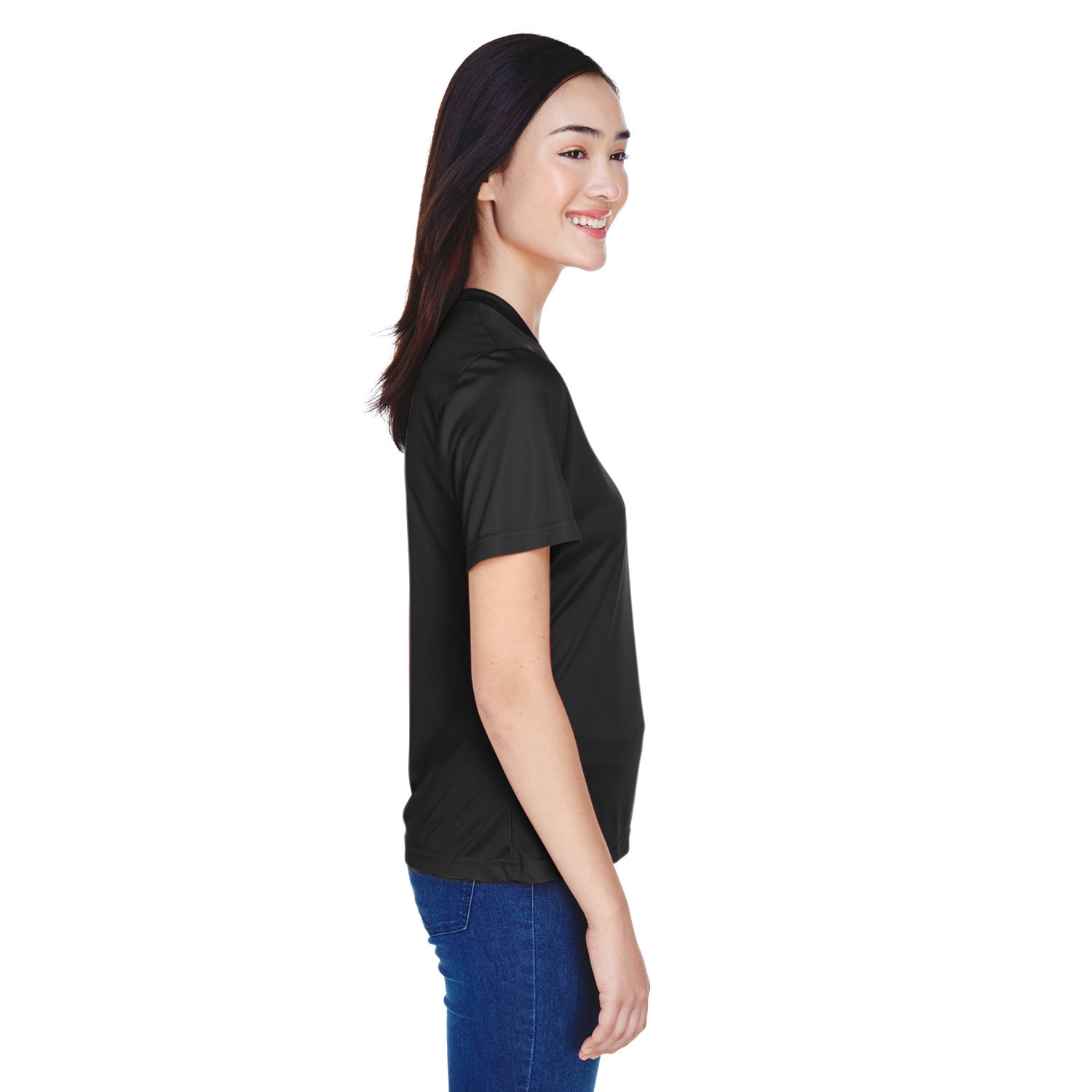 Smiling woman in black short-sleeve shirt and blue jeans standing in profile against white background