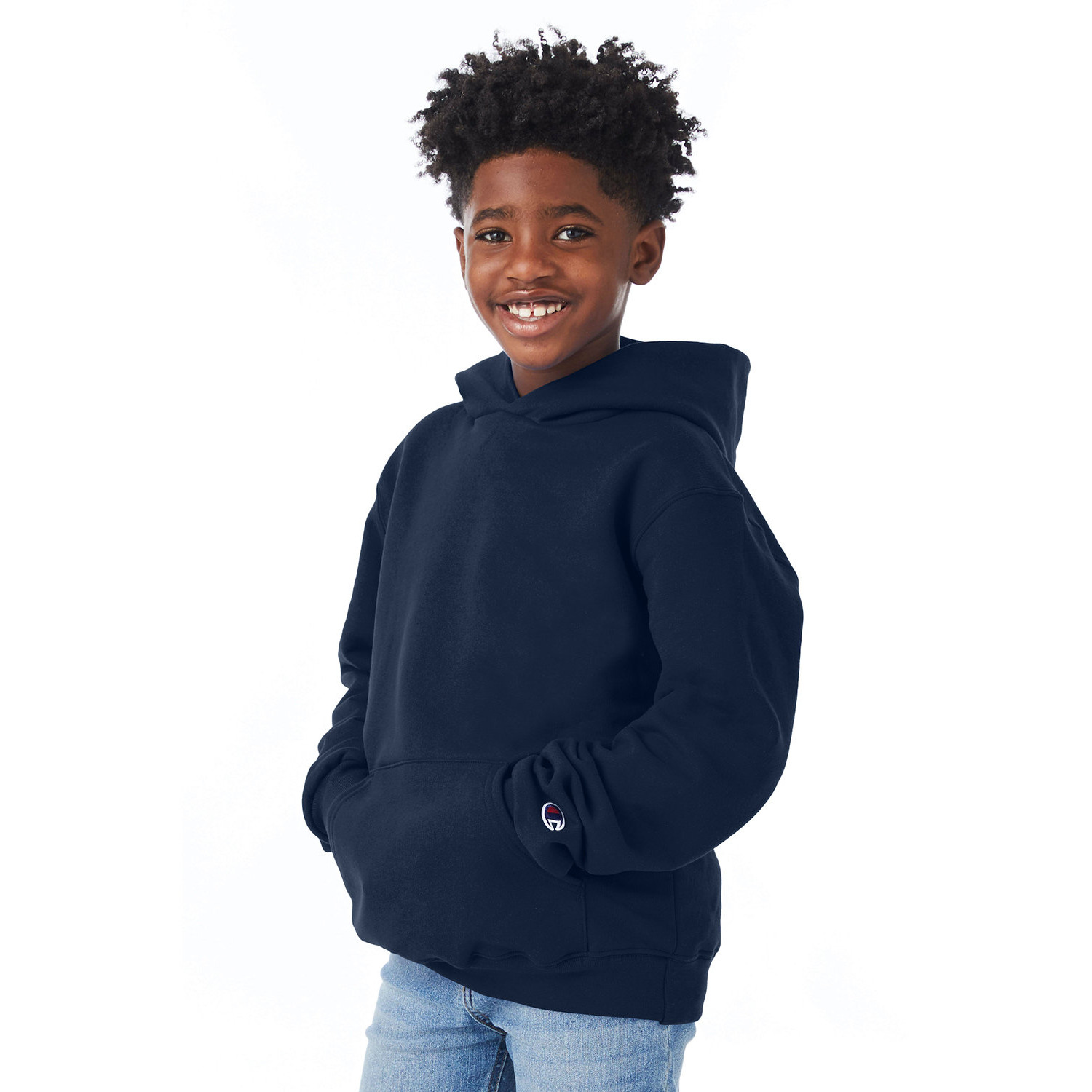 Smiling young boy with curly hair wearing a navy blue hoodie and light blue jeans standing with hands in hoodie pocket against a white background