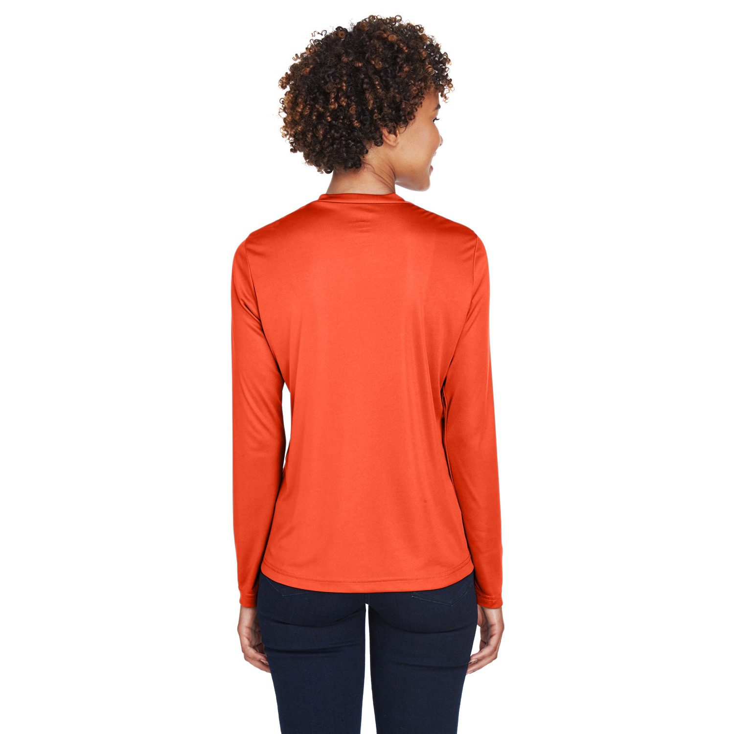 team_365_zone_ladies_ls_back Rear view of a person with curly hair wearing an orange long-sleeve shirt and dark blue pants against a white background.
