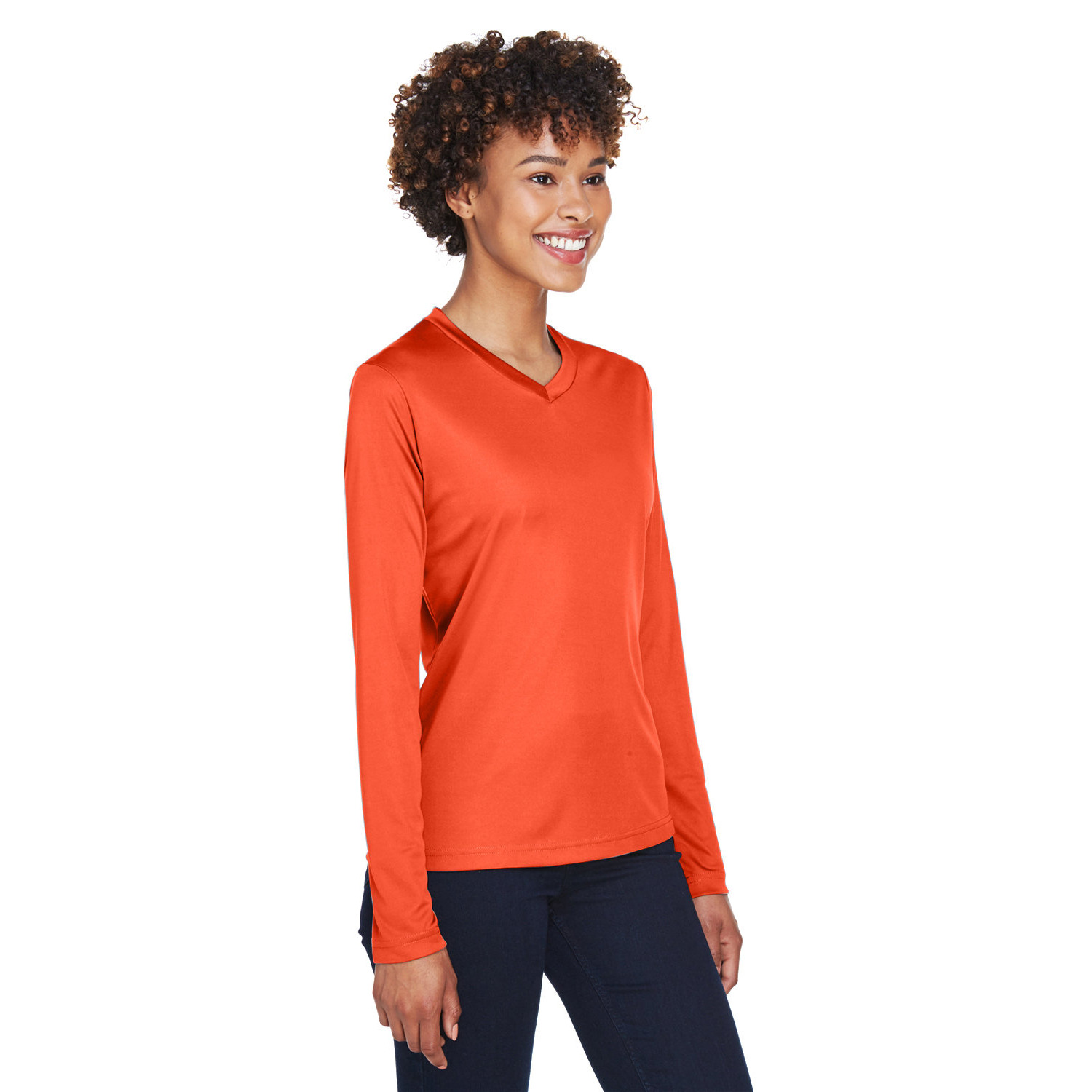 team_365_zone_ladies_ls_front Smiling woman with curly hair wearing a bright orange long-sleeve V-neck shirt and dark pants against a white background