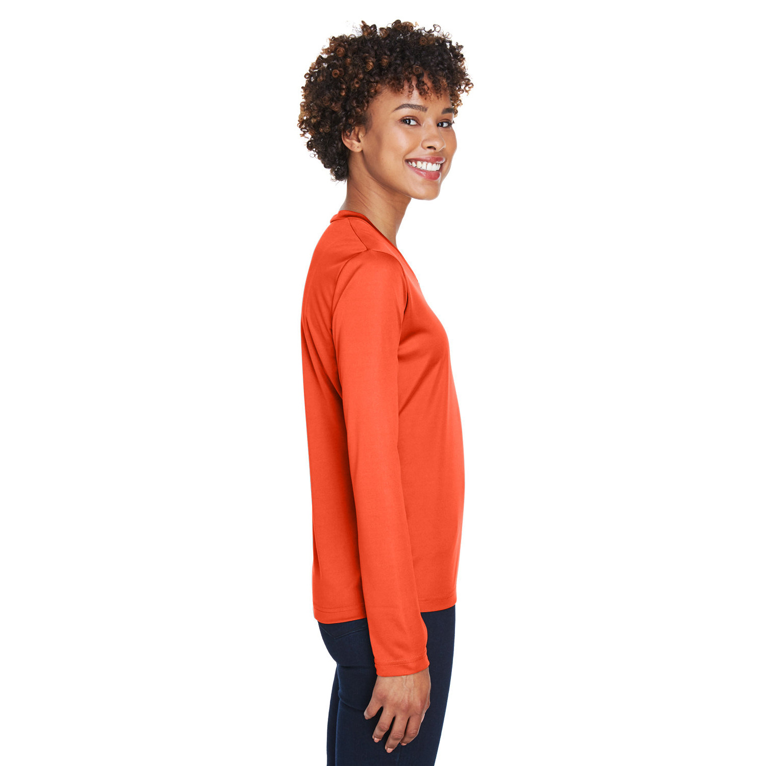 team_365_zone_ladies_ls_side Young woman with curly hair wearing an orange long-sleeve shirt and dark pants, smiling and standing in profile against a white background