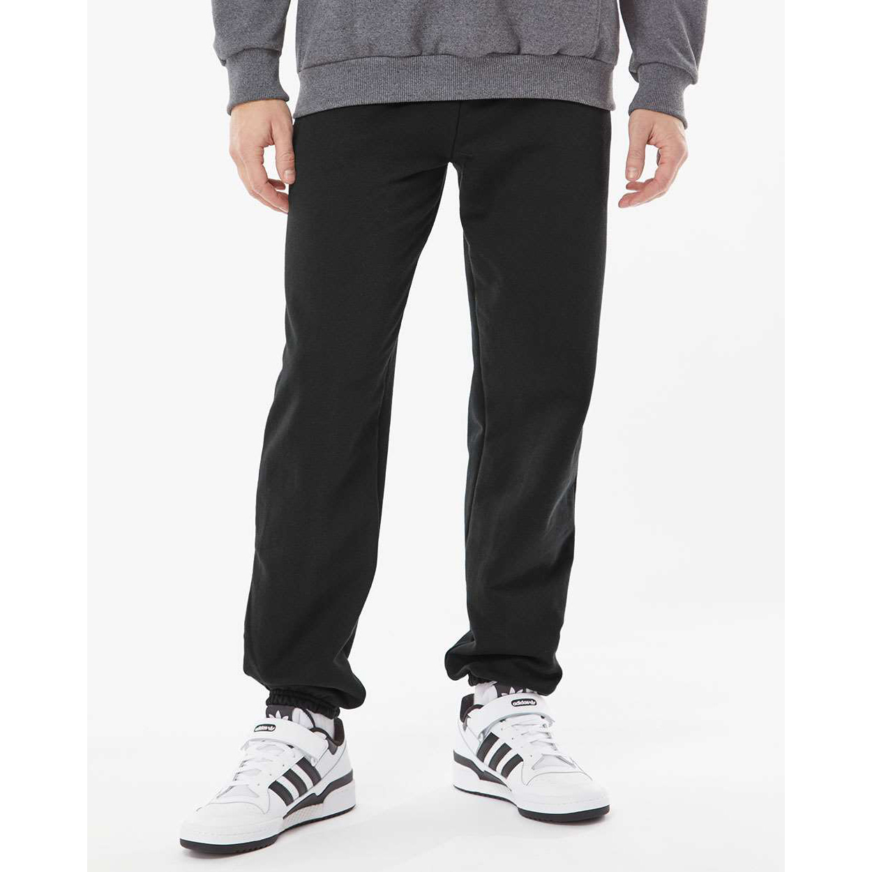 Lower half of a person wearing black sweatpants and white Adidas sneakers with black stripes, standing against a white background.
