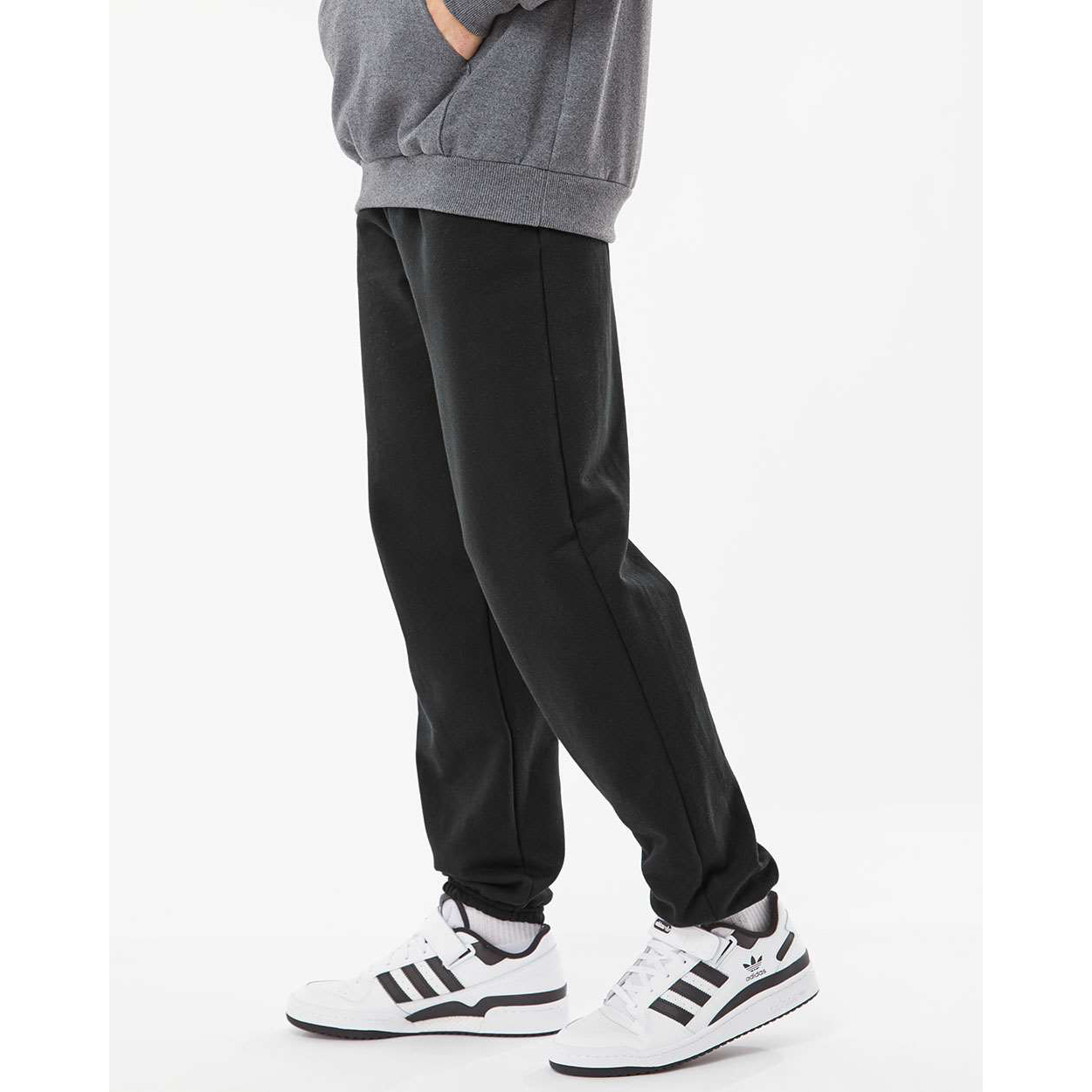 Lower body of a person wearing black sweatpants, white Adidas sneakers with black stripes, white socks, and a gray sweatshirt with hands in pockets against a plain white background.