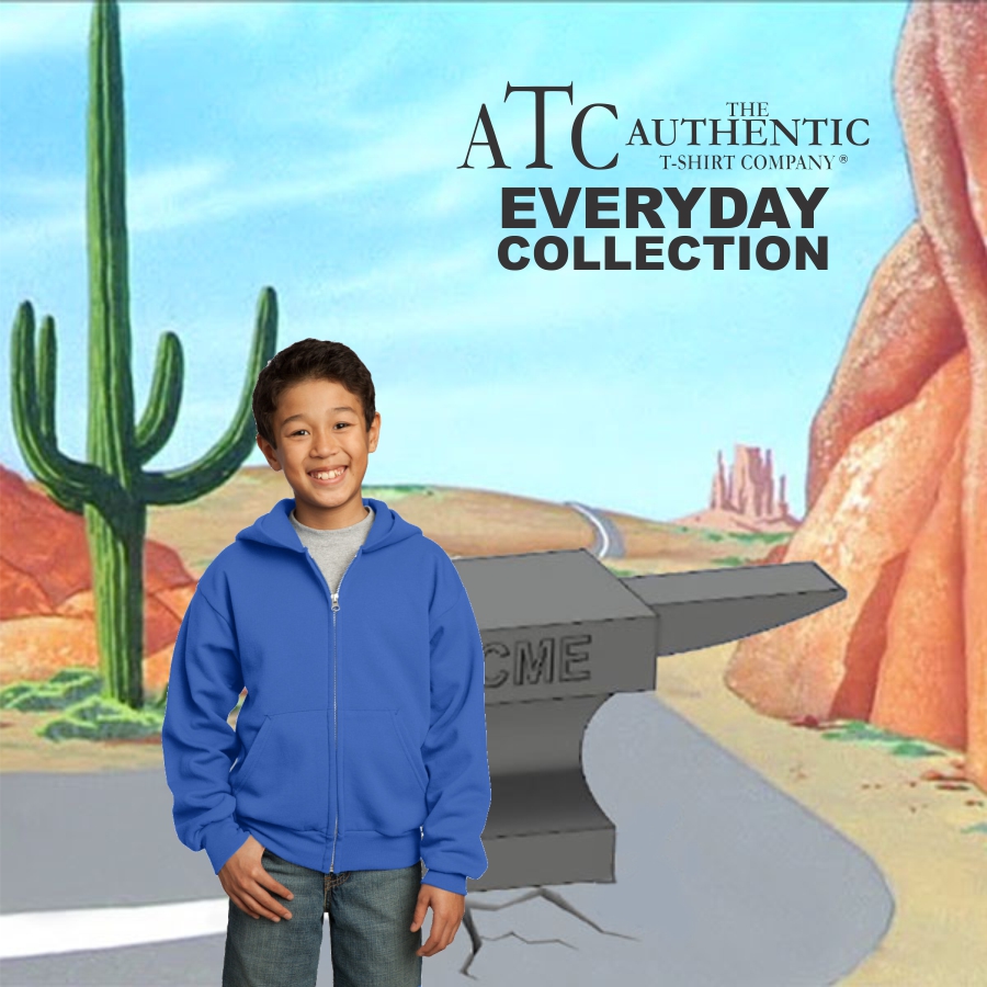 A smiling boy wearing a blue zip-up hoodie and jeans standing in front of a cartoon desert road scene featuring an anvil labeled "ACME" and a large cactus, with text above reading "ATC The Authentic T-Shirt Company Everyday Collection".