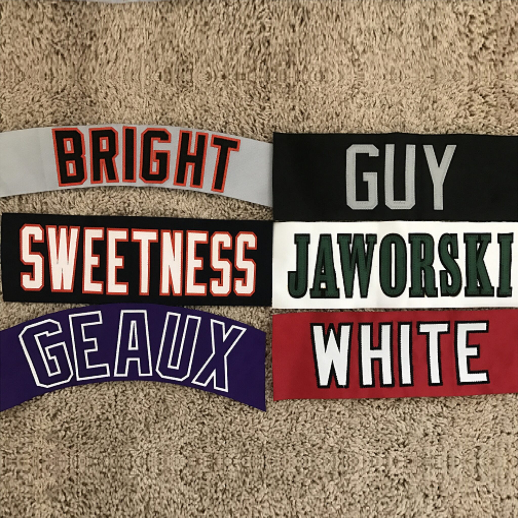 Six fabric patches with words in block letters on a textured beige surface, reading BRIGHT in red and black on gray, GUY in gray on black, SWEETNESS in white and orange on black, JAWORSKI in green and black on white, GEAUX in white on purple, and WHITE in white and black on red.