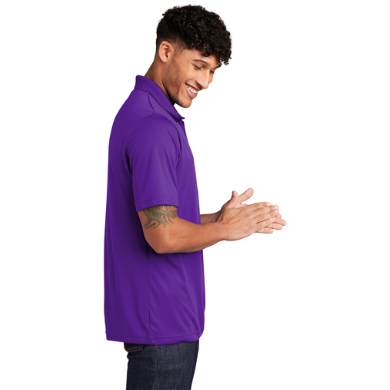 Side view of a smiling man with curly hair wearing a purple polo shirt and dark jeans, clapping his hands together against a white background.