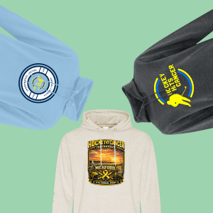 Three hoodies featuring designs for the "Hockey Hits Cancer" tournament, including a blue hoodie with a circular logo, a beige hoodie with a colorful event graphic, and a dark gray hoodie with bold yellow text, all set against a light green background.